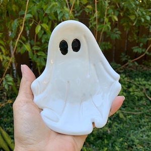 May include: A white ceramic trinket dish shaped like a ghost with a smooth, glossy finish. The ghost figure features two simple, oval-shaped black painted eyes centered near the top, with molded folds and curves representing a draped sheet.