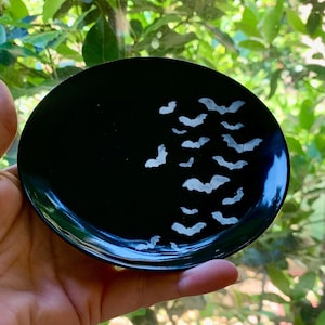 May include: A small, round, black dish with a glossy finish. The dish is decorated with a cluster of white bat silhouettes. The bats are arranged in a descending pattern across the surface. The dish is held in a hand.