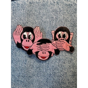 Könnte beinhalten: Gestickter Aufnäher mit drei Affen in der klassischen Pose „nichts sehen, nichts hören, nichts sagen“. Die Affen haben schwarze Gesichter, rosa Hände und Ohren sowie rote Münder. Auf blauem Jeansstoff.