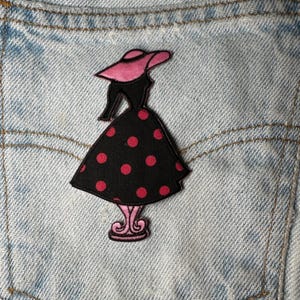 Puede incluir: Parche bordado en negro y rosa con un vestido y sombrero con lunares. El parche está sobre un fondo de mezclilla.