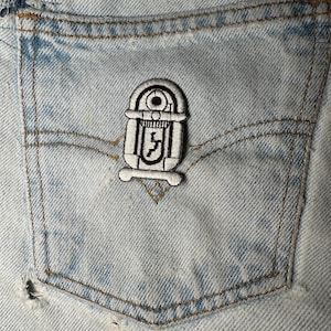 Puede incluir: Un bolsillo de jean de mezclilla azul claro con un pequeño parche bordado. El parche es un diseño de gramola en blanco y negro. El bolsillo tiene costuras visibles y un pequeño desgarro en la parte inferior.