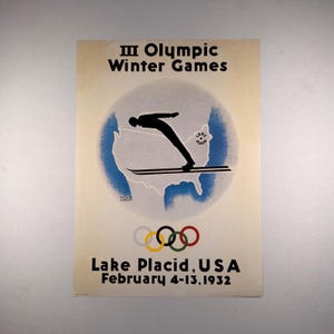 May include: A vintage poster for the III Olympic Winter Games in Lake Placid, USA, February 4-13, 1932. It features a black silhouette of a ski jumper over a blue map of the United States, with the Olympic rings and bold black text.