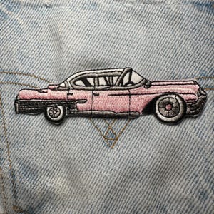 May include: Pink embroidered patch of a classic car with black and white detailing. The patch is rectangular and features a detailed design of the car's body, wheels, and windows. The patch is sewn onto a light blue denim fabric.