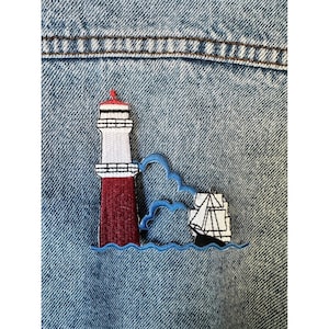 May include: Embroidered patch featuring a red and white lighthouse with a red top, a sailboat, and blue waves. The patch is sewn onto a blue denim jacket.