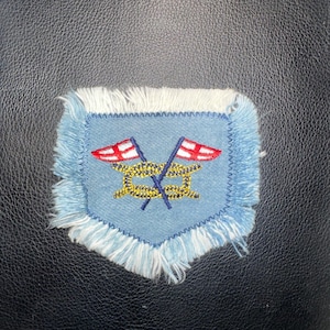 May include: A light blue embroidered patch with a shield shape. The patch features two crossed flags with red and white designs, and a gold-colored knot. The edges are fringed with white and light blue threads.