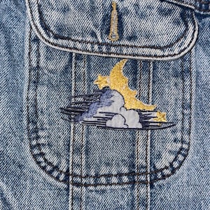 Puede incluir: Una chaqueta vaquera azul con un parche bordado en oro y azul de una luna creciente y nubes en el bolsillo.