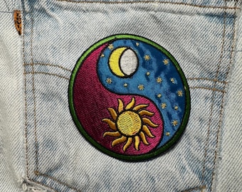 May include: Embroidered yin and yang patch featuring a sun, moon, and stars design. The patch has a green border and is sewn onto the back pocket of a light-wash denim garment.