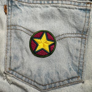 May include: Close-up of a light blue denim pocket with a circular patch. The patch features a yellow star inside a red circle, outlined in green and yellow. The denim shows wear and tear.
