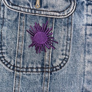 May include: Close-up of a light blue denim jacket pocket with a purple embroidered sunburst design. The jacket has visible stitching and a button. The overall theme is casual wear.