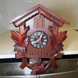 May include: A dark brown wooden cuckoo clock featuring a gabled roof, carved leaf accents on the sides, and a bird figure at the bottom. The clock face has a black background with white Roman numerals and the word GERMANY printed on it.