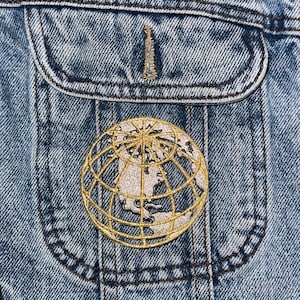 May include: A close-up of a denim jacket with a gold embroidered globe patch on the pocket.