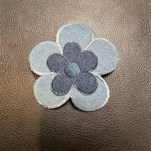 May include: A denim flower patch with a light blue and dark blue color scheme. The flower has five petals and a circular center. The patch is likely for clothing or accessories.