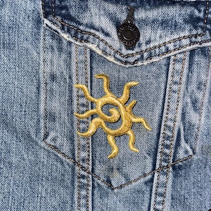 May include: A blue denim jacket with a gold embroidered sun design on the pocket flap.