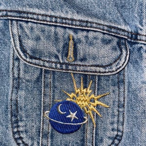 Può includere: Una giacca di jeans blu con una toppa ricamata in oro di un pianeta con una luna crescente e una stella, circondata da un sole con raggi.