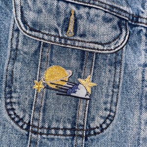 Puede incluir: Una chaqueta vaquera azul con un bolsillo que presenta un parche bordado de un planeta amarillo, una estrella fugaz y una nube.