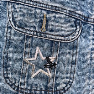 May include: A close-up of a blue denim jacket with a silver star patch on the pocket. The star has a black outline and is surrounded by smaller black stars.