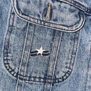 May include: Close-up of a blue denim jacket with a pocket featuring a white star and two black stripes.