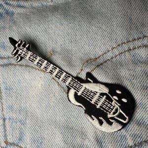 Pode incluir: Um patch de guitarra bordado em preto e branco. O patch apresenta um design detalhado de uma guitarra elétrica com costuras brancas delineando o corpo e a escala. O patch é colocado em um fundo de ganga azul claro.