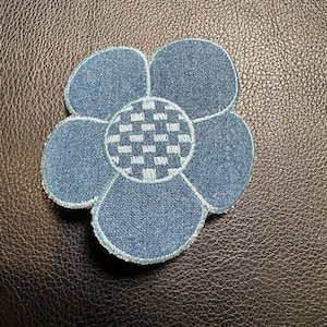May include: A blue denim flower appliqué with five petals and a checkered centre. The patch has white stitching and is set against a dark brown textured background.