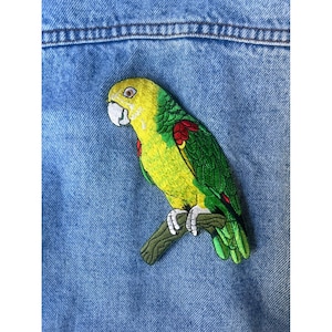 May include: Embroidered parrot patch featuring a yellow head, green body, and red accents. The parrot is perched on a brown branch against a blue denim background. The patch is likely for clothing or accessories.