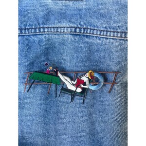May include: Embroidered patch on a blue denim jacket. The patch depicts a woman in a red swimsuit reclining on a chair with a drink and a life preserver. The woman is wearing a yellow hat and is on a wooden structure.