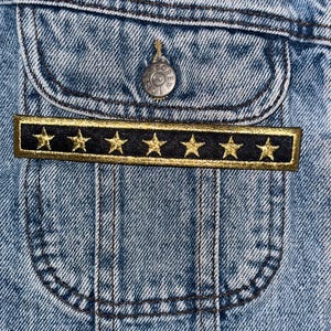 May include: A close-up of a denim jacket pocket with a black and gold patch featuring five gold stars. The patch is sewn onto the pocket flap.