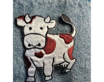 May include: A white and red cow patch with black stitching, set against a blue denim background. The cow has red spots and a black tail. This embroidered patch is a fun accessory for jackets, bags, or other fabric items.