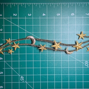 Puede incluir: Parche bordado en oro y plata de una luna creciente y estrellas en línea curva sobre un fondo de cuadrícula verde.