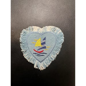 May include: A heart-shaped patch with a light blue background and a sailboat design. The sailboat has yellow and blue sails, a red hull, and is set against a blue water background. The patch is trimmed with a fringe.