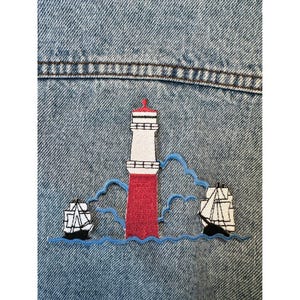 May include: Embroidered patch of a red and white lighthouse with two sailing ships on blue waves, sewn onto a denim jacket. The lighthouse is positioned between the ships.