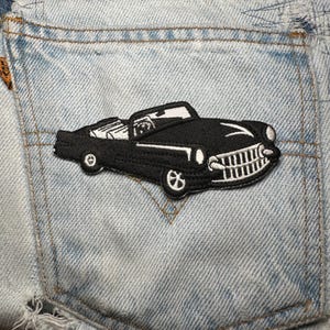 May include: Black embroidered patch of a classic convertible car with white detailing. The patch is affixed to a light blue denim pocket. The car features a detailed grill, headlights, and wheels, with a white windshield frame.