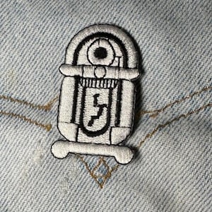 May include: An embroidered patch featuring a white and black jukebox design with a rounded top, a circular speaker detail, and a central vertical panel with a jagged black line graphic, all outlined in thick black thread on a light blue denim fabric.