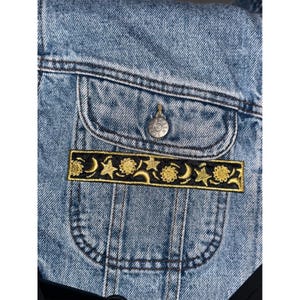 May include: A light blue denim jacket with a pocket featuring a black and gold embroidered patch. The patch displays a celestial design with moons, stars, and suns. The jacket has a button closure and a classic design.