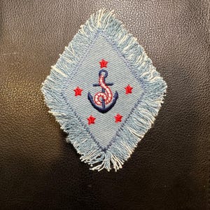 Może przedstawiać: Plaster w kształcie rombu z jasnoniebieskim dżinsowym tłem i białymi frędzlami. Na naszywce znajduje się granatowa kotwica z czerwoną liną i trzema czerwonymi gwiazdami. Tematyka żeglarska.