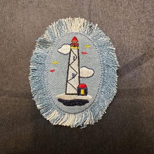 May include: Oval-shaped denim coaster with a lighthouse design. The lighthouse is white with a red top and sits on a small island. The coaster has a light blue fringe and includes embroidered clouds and birds.