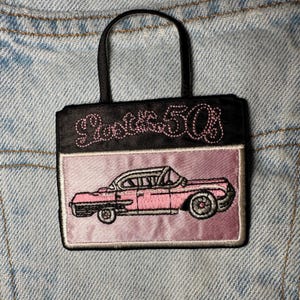 以下が含まれることがあります： ピンク色の刺繍された車と、ピンク色のスクリプトで「Lost in the 50s」の文字が入った、ハンドルの付いた小さな黒いハンドバッグ。 バッグは長方形で、上部は黒、下部はピンクです。