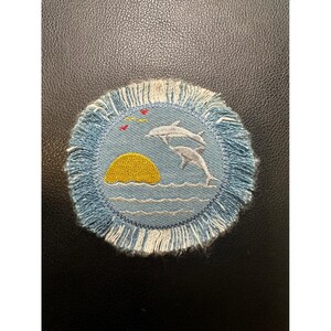 May include: Embroidered circular patch featuring a sunset over ocean waves with two leaping dolphins. The patch has a light blue background with a yellow sun and white dolphins. The border is fringed with light blue and white threads.