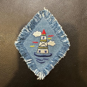 May include: A diamond-shaped denim patch with a fringed edge. The patch features an embroidered lighthouse with a red roof, white body, and blue base, set against a blue sky with clouds and red and yellow accents. The lighthouse is surrounded by blue water.