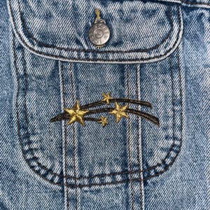 以下が含まれることがあります： 青いデニムジャケットのアップで、ポケットに金色の刺繍で描かれた流れ星のデザインが施されています。