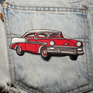 May include: Embroidered patch of a red classic car with white detailing, attached to a light blue denim pocket. The car has black tyres and a silver grille, with a detailed design. The patch is a decorative item.