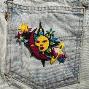 May include: Light blue denim pocket with a colorful embroidered patch. The patch features a yellow sun, a maroon crescent moon, stars, and a planet with a yellow ring. The patch is stitched onto the pocket.