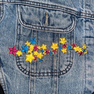 May include: A blue denim jacket with a pocket on the front. The pocket has a colorful embroidered design of stars in yellow, pink, and blue.