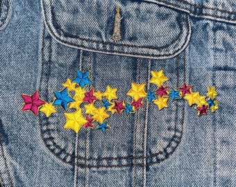 May include: A blue denim jacket with a pocket on the front. The pocket has a colorful embroidered design of stars in yellow, pink, and blue.