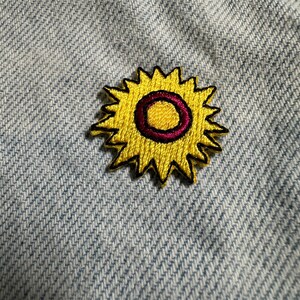 Könnte beinhalten: Gestickter Aufnäher mit einer gelben Sonnenblume mit dunkelviolettem Zentrum und schwarzer Umrandung. Der Aufnäher ist auf hellblauem Denim-Stoff aufgenäht, wodurch ein Kontrast in Texturen und Farben entsteht. Das Sonnenblumen-Design ist ein fröhliches, dekoratives Element.