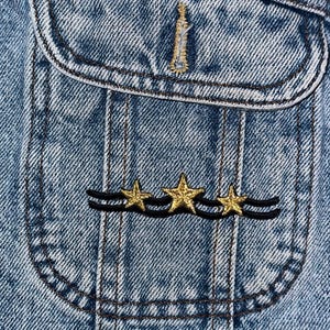 Puede incluir: Primer plano de un bolsillo de chaqueta vaquera con un bordado de estrella dorada y ola negra.