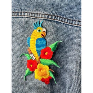 Könnte beinhalten: Gestickter Aufnäher mit einem bunten Papagei, der auf einem Ast mit roten und gelben Blumen und grünen Blättern sitzt. Der Papagei hat gelbe, blaue und weiße Federn. Der Aufnäher ist auf einer Jeansjacke befestigt.