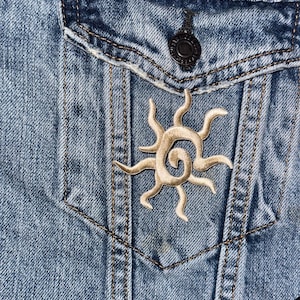 May include: Close-up of a light blue denim jacket with a pocket and a decorative beige sun patch. The jacket has a black button and black trim.