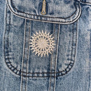 May include: Close-up of a denim jacket with two decorative pins. One pin is a sunburst design, and the other is a small, vertical pin. The jacket has a classic pocket design and is made of blue denim fabric.