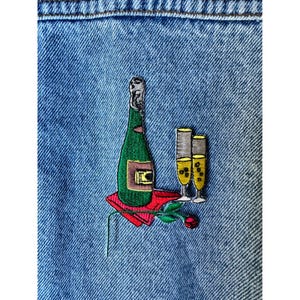 May include: Embroidered patch featuring a green champagne bottle, two champagne flutes, and a red rose on a denim background. The bottle has a gold label, and the flutes are filled with yellow liquid.