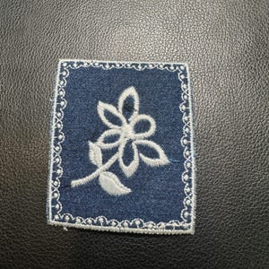 May include: A square, navy blue patch with a white floral design. The flower is embroidered with delicate details, and the patch is bordered with a white, scalloped edge. The patch is likely for clothing or accessories.